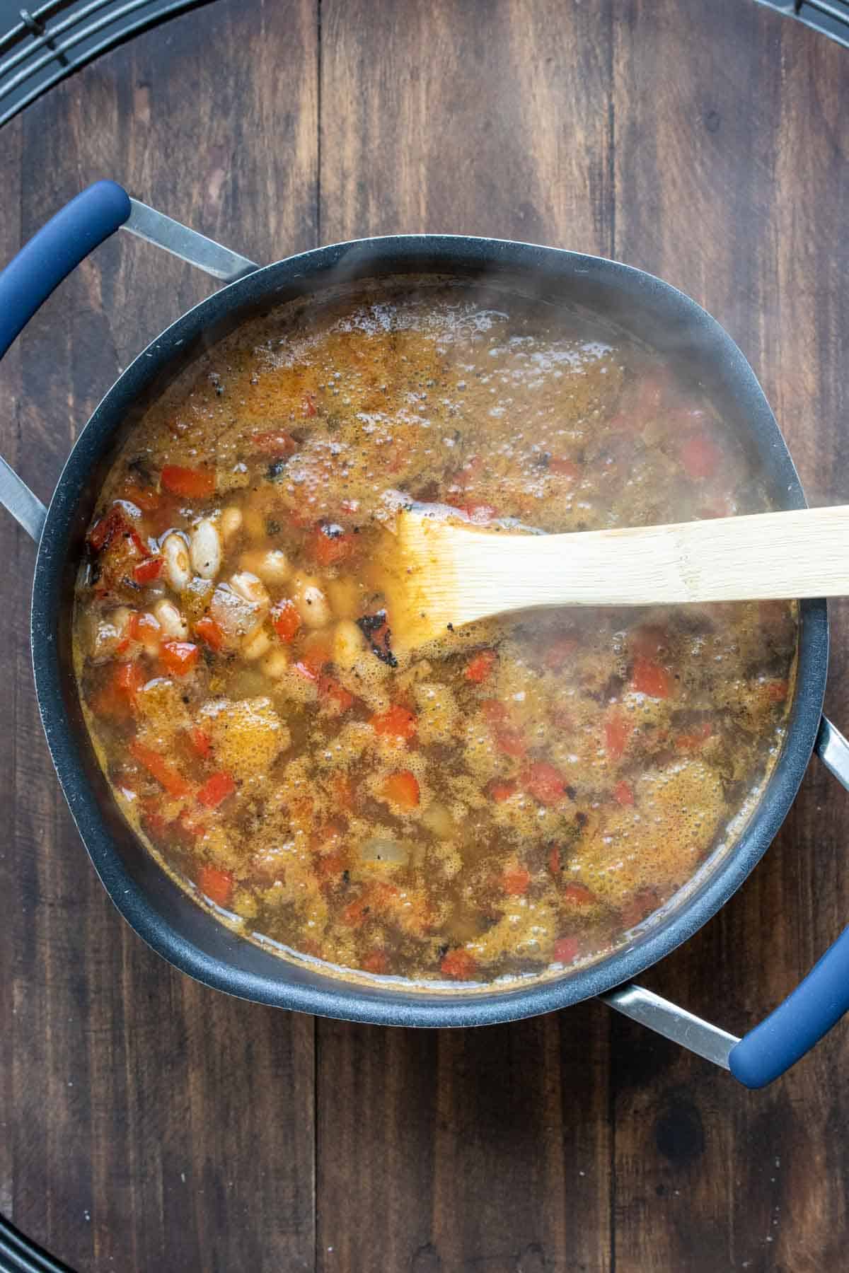 Wooden spoon mixing a pot with broth, dried pinto beans and red pepper pieces