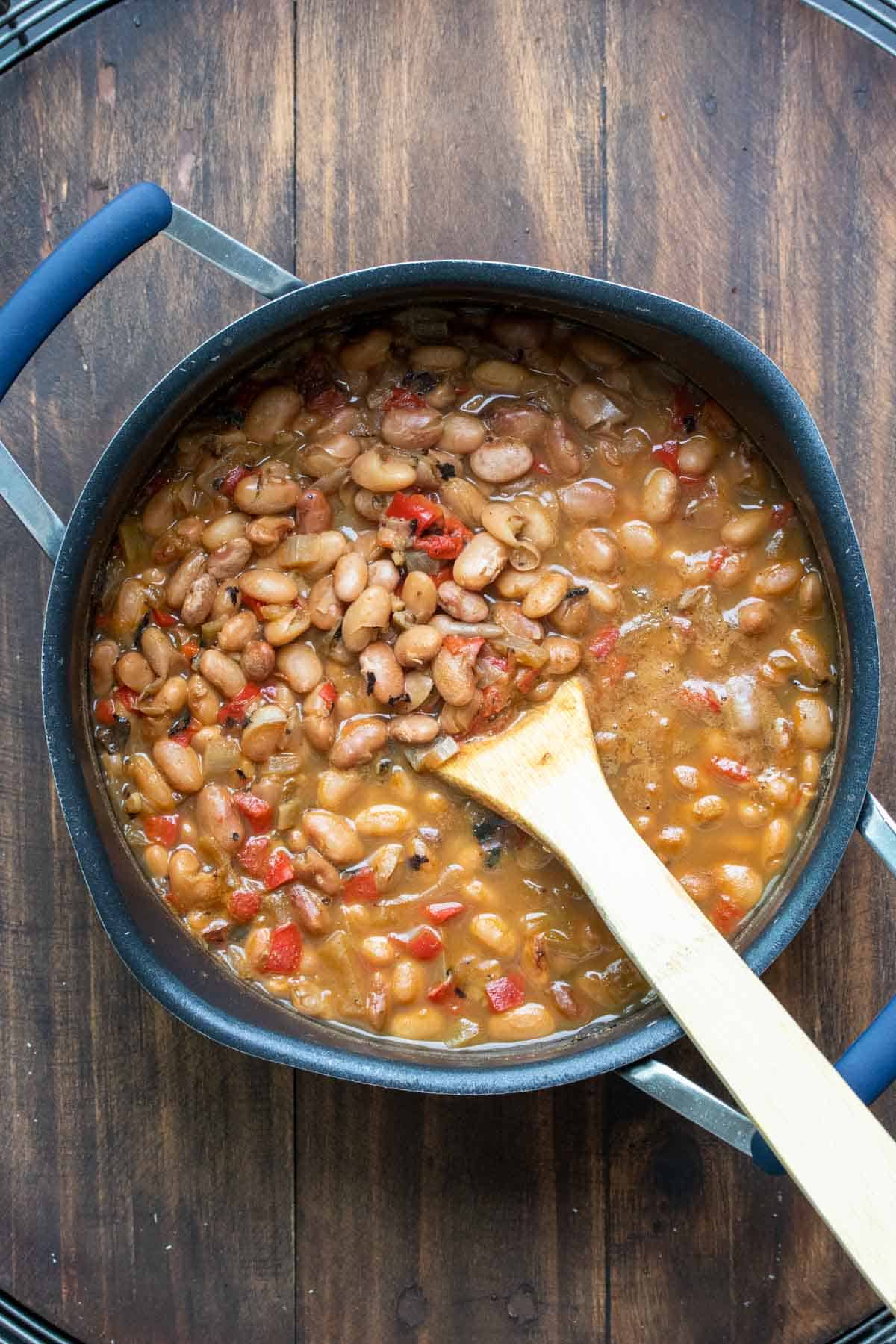 Wooden spoon mixing a pot of liquidy pinto beans with chopped red pepper