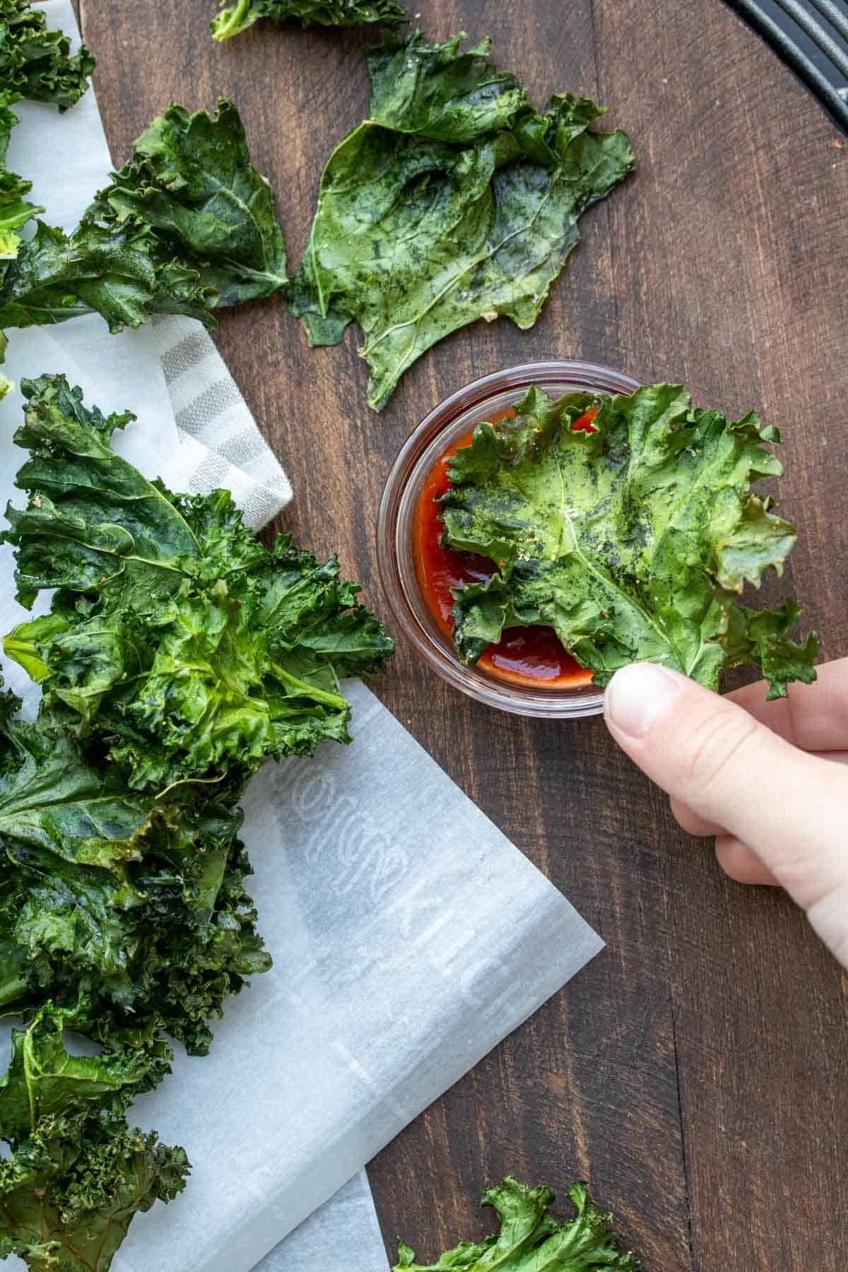 Hand dipping a kale chip into a glass dish of ketchup