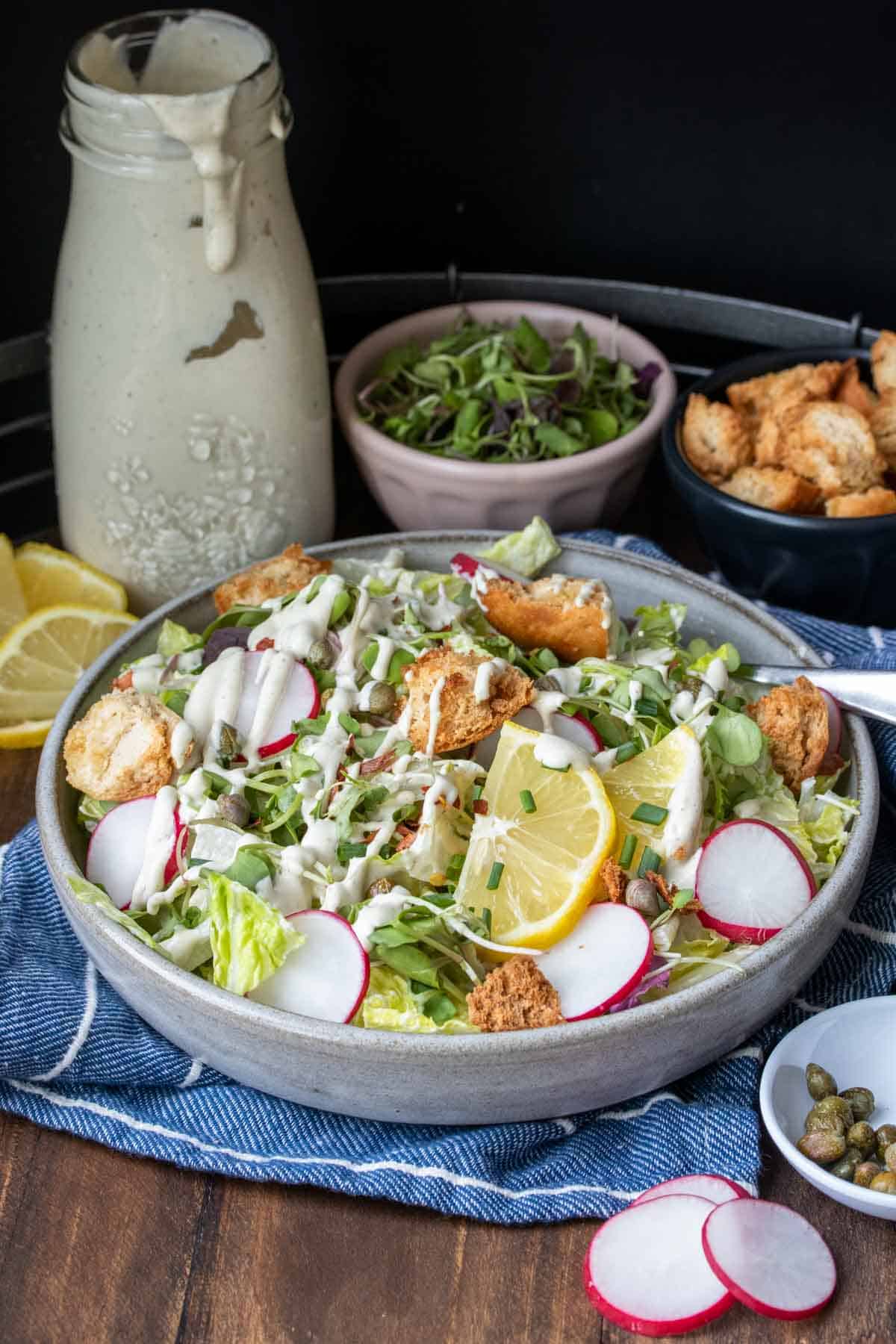 Grey plate with a dressed salad in front of bowls of toppings and dressing