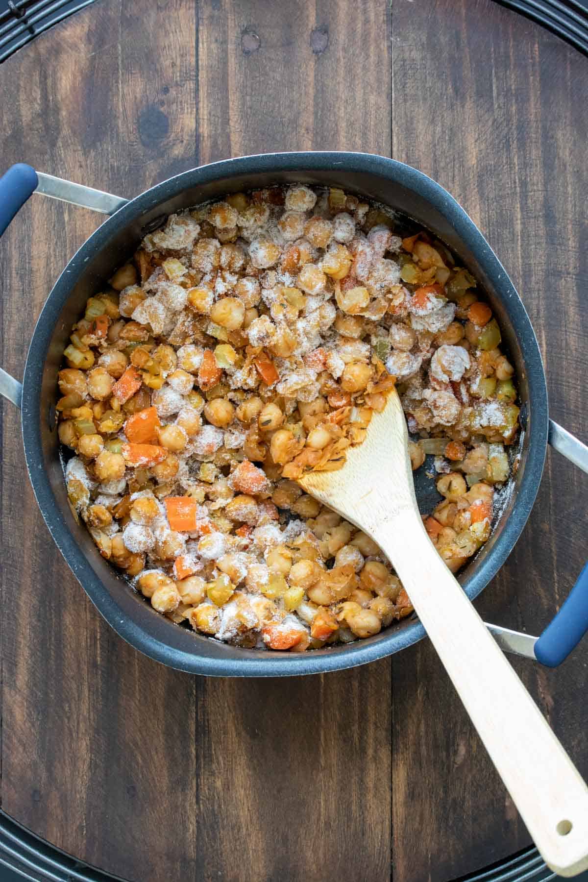 Wooden spoon mixing veggies and chickpeas topped with flour in a pot.