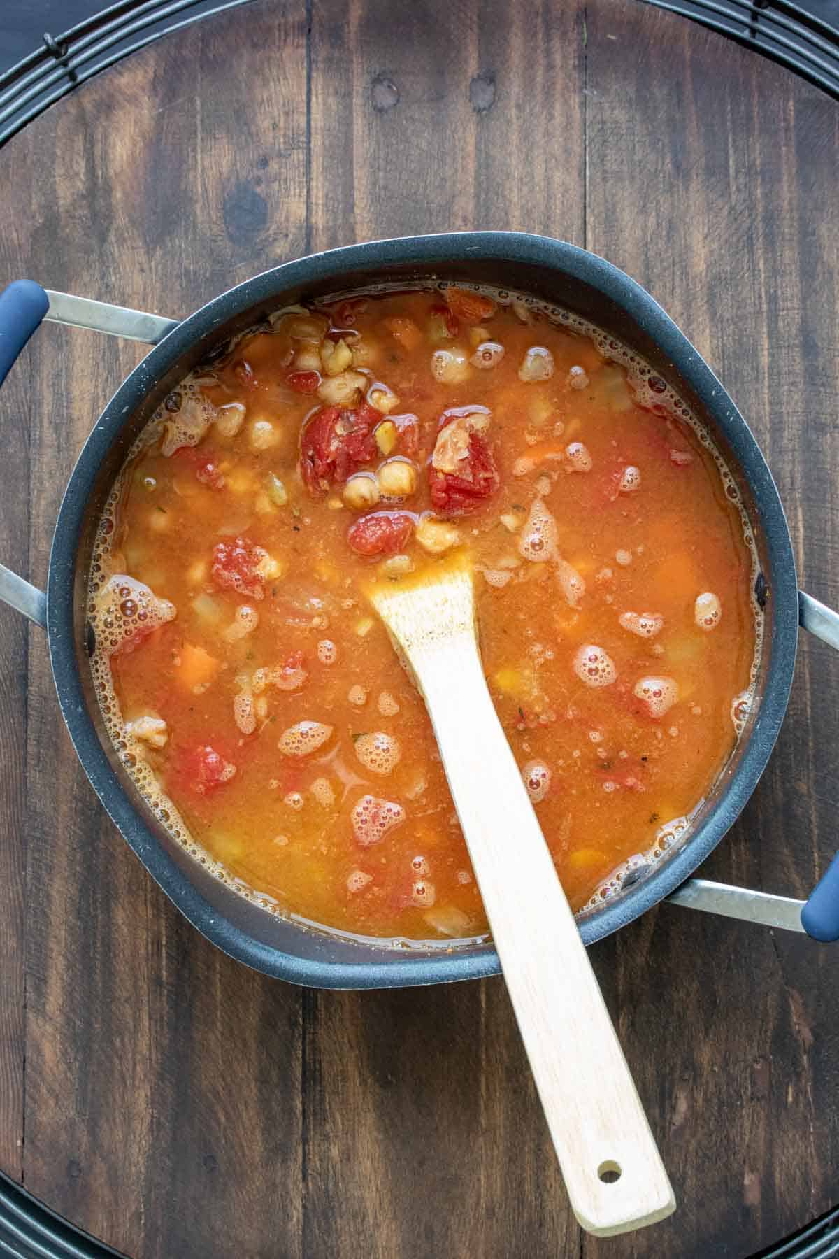 Pot with broth, tomatoes and chickpeas being mixed by a wooden spoon.