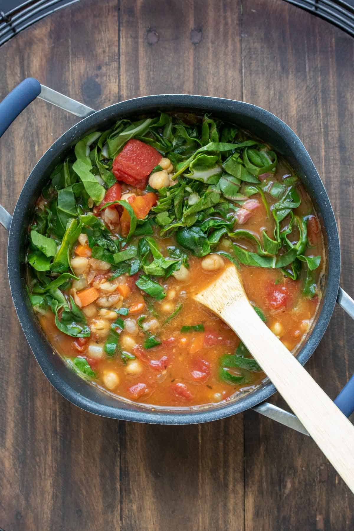 Wooden spoon mixing greens into a pot with broth, chickpeas and tomatoes.