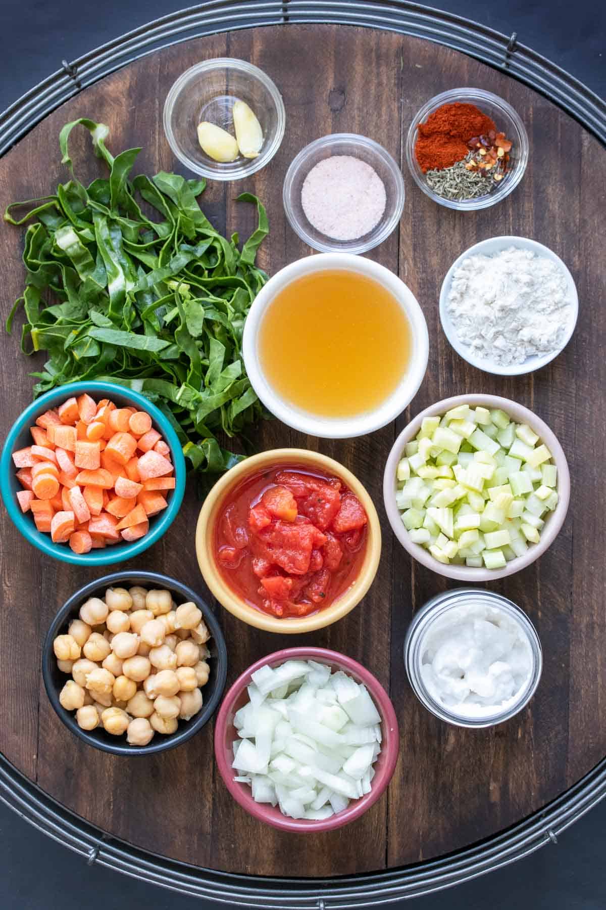 A wooden table covered in bowls with veggies, chickpeas, broth and spices.