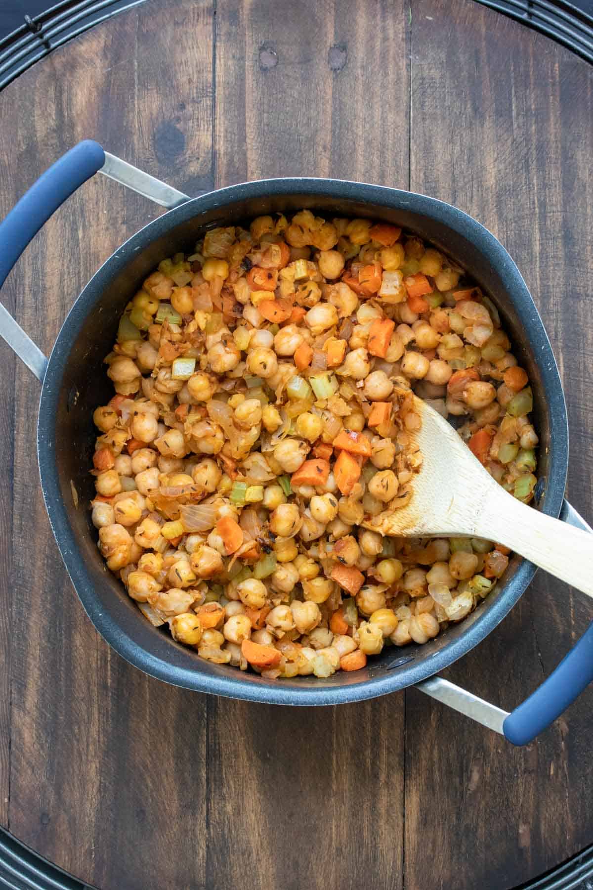 Wooden spoon mixing veggies and chickpeas with spices in a pot.