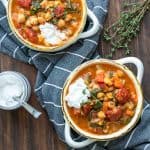 Two bowls filled with veggie and chickpea stew on a wooden table next to a jar of yogurt.