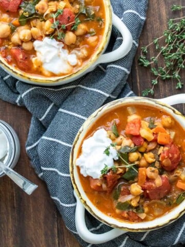 Two bowls filled with veggie and chickpea stew on a wooden table next to a jar of yogurt.