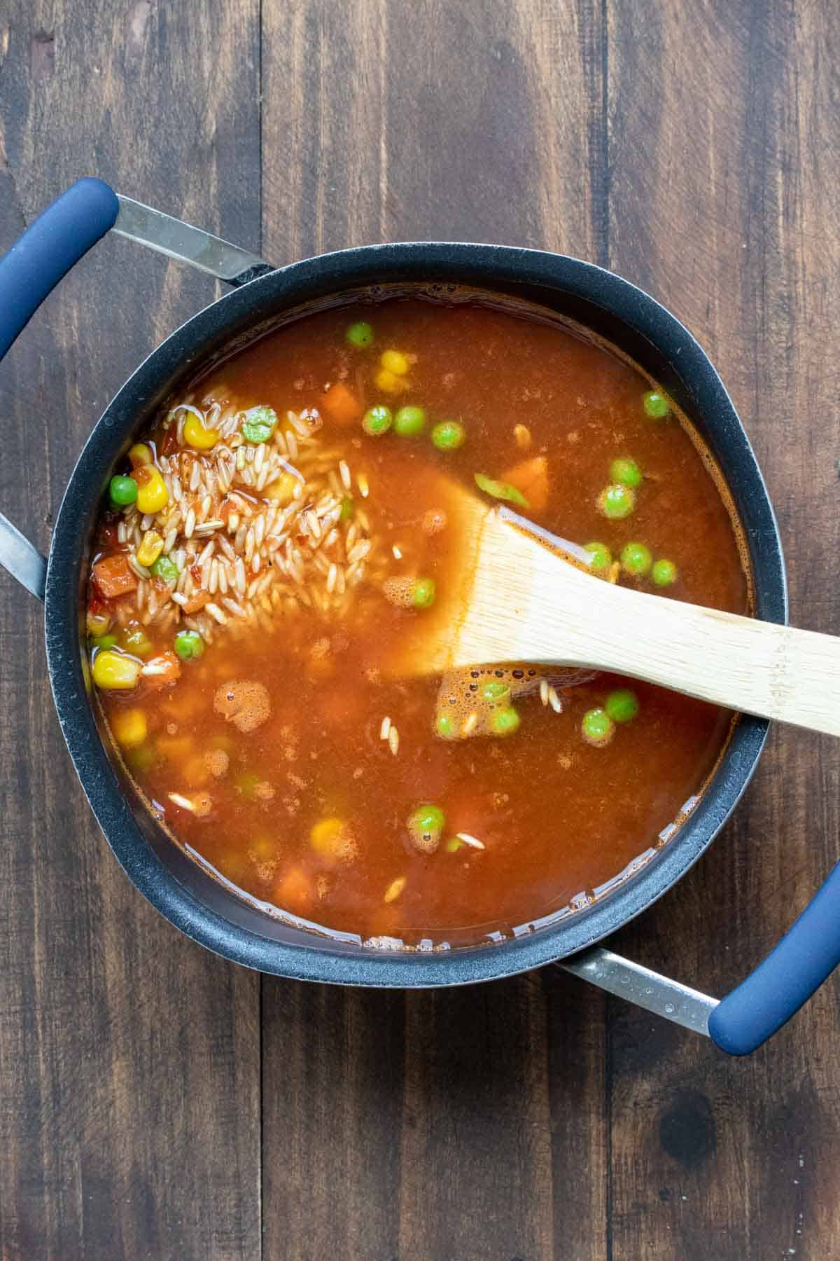 Wooden spoon mixing broth, rice, peas and carrots in a pot