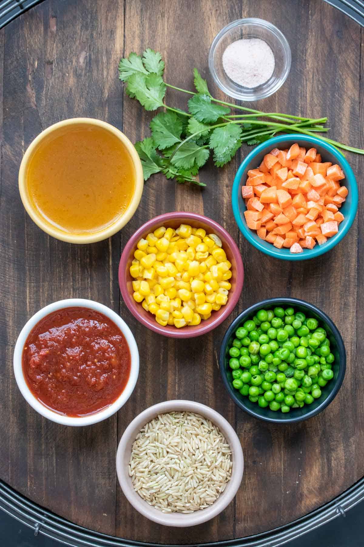 Bowls with rice, salsa, carrots, corn, peas and broth next to cilantro and salt