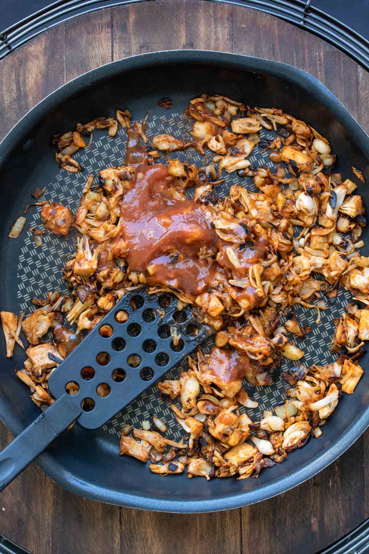 Spatula mixing BBQ sauce over pieces of browned jackfruit in a pan.