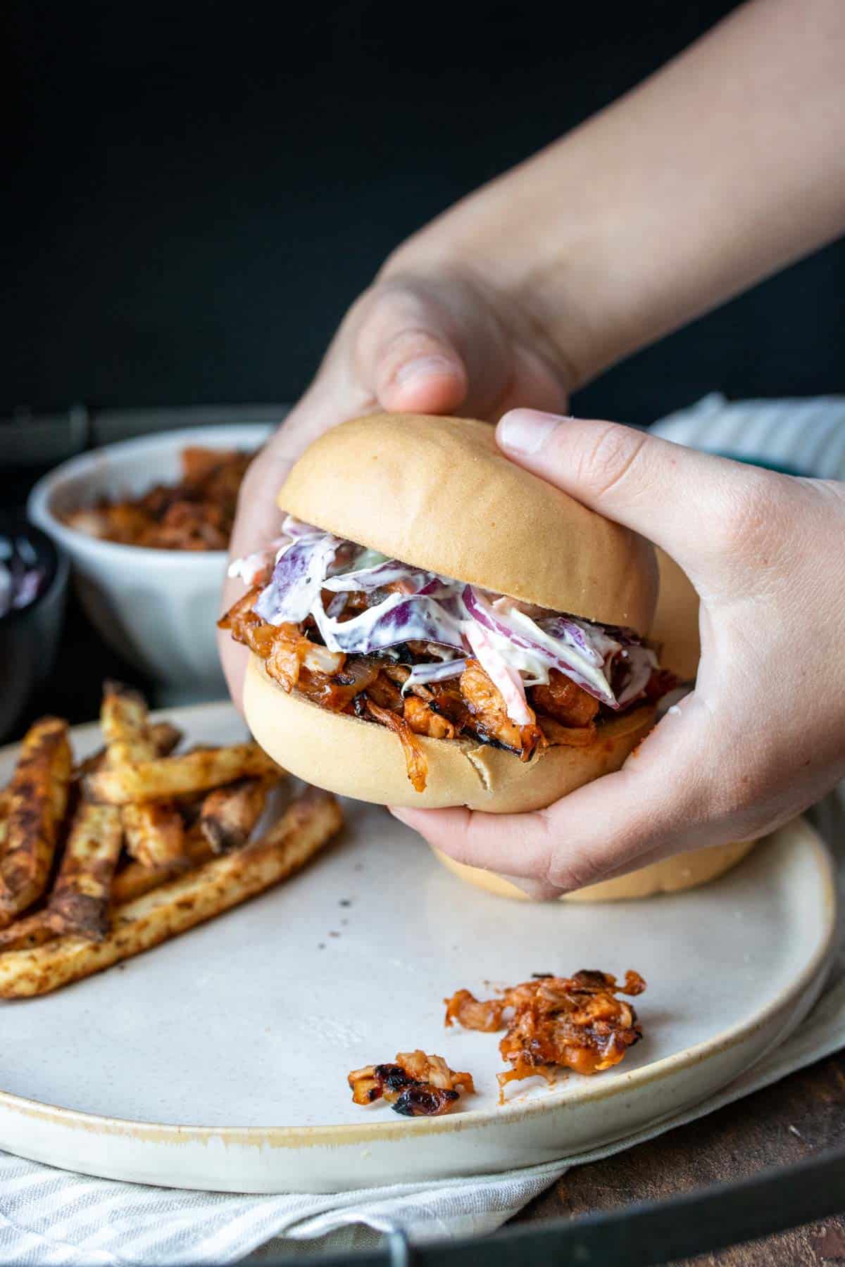Hands picking up a BBQ jackfruit sandwich topped with coleslaw off a plate