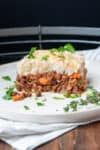 A piece of cottage pie with a vegetarian filling sitting on a white plate with parsley on and around it.