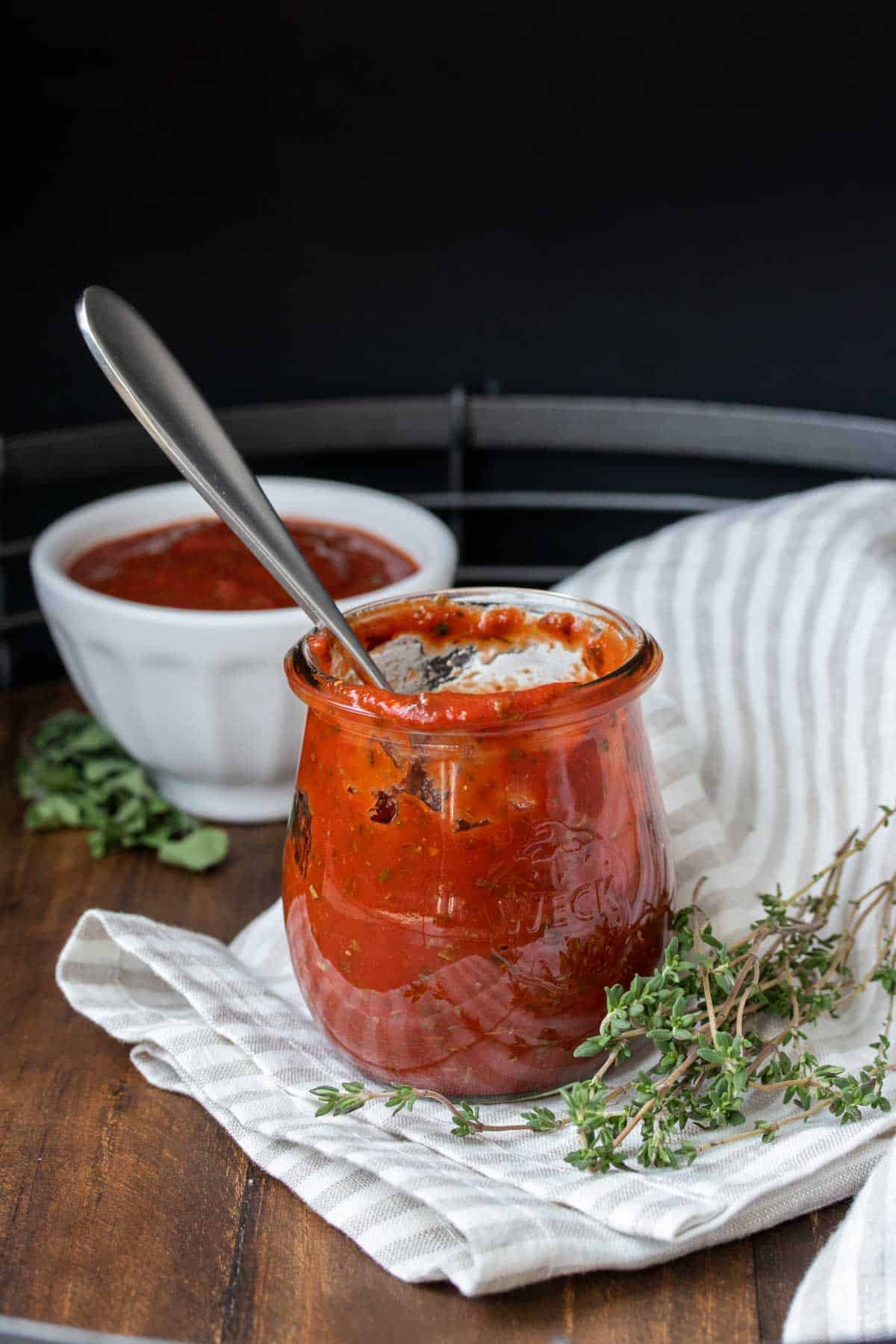 A glass jar on a striped towel with a spoon and pizza sauce inside