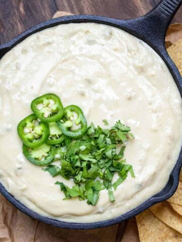 A black cast iron pan filled with white queso dip that is topped with sliced jalapenos and chopped cilantro.