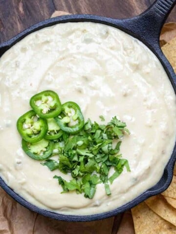 A black cast iron pan filled with white queso dip that is topped with sliced jalapenos and chopped cilantro.