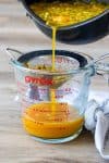 A pot pouring passion fruit syrup through a strainer into a glass cup