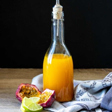 A glass bottle filled with orange liquid sitting on a towel next to limes and passion fruit