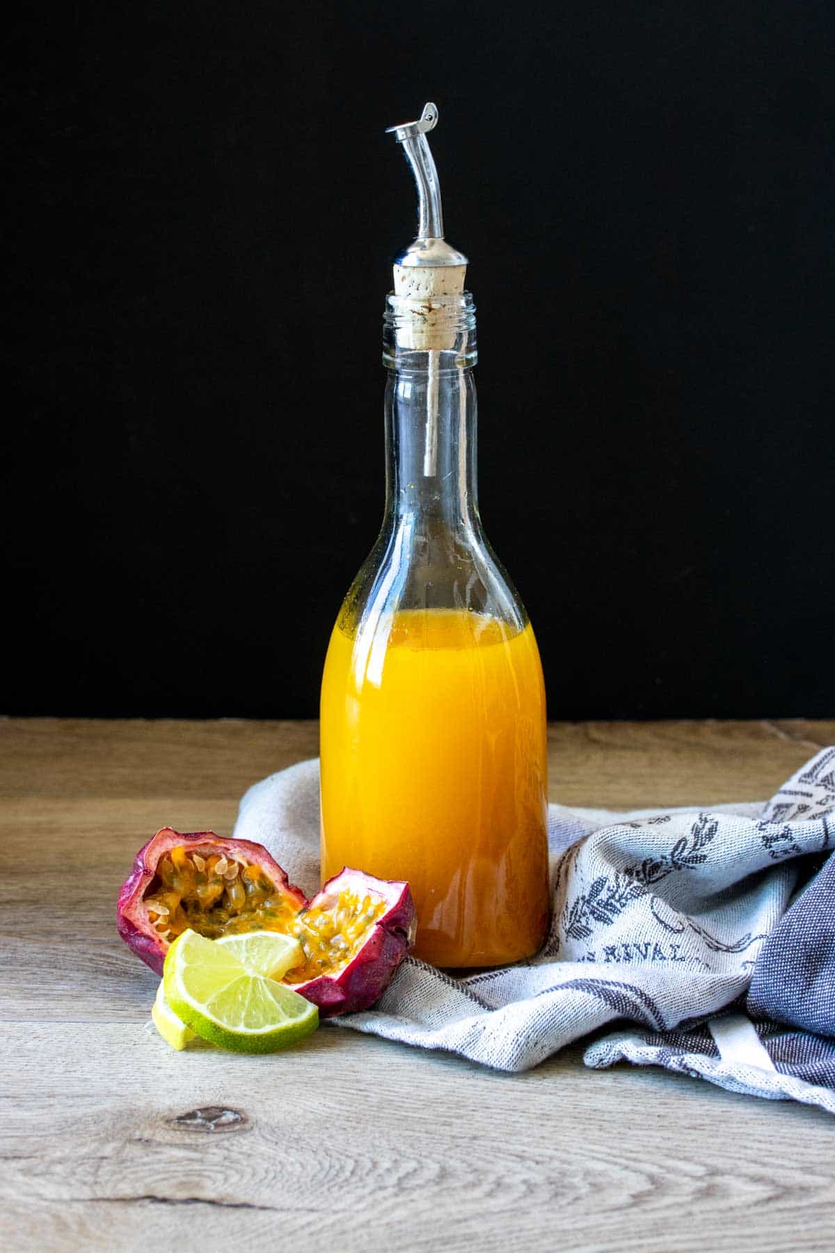 A glass bottle filled with orange liquid sitting on a towel next to limes and passion fruit