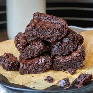 A pile of brownies on parchment paper with a glass of milk behind.