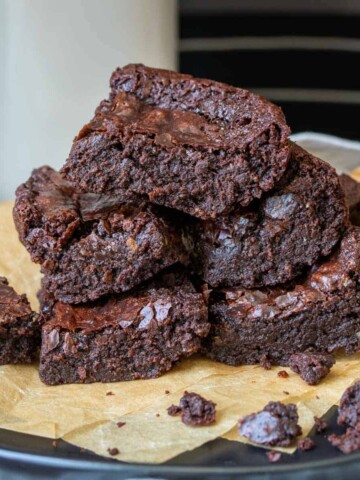A pile of brownies on parchment paper with a glass of milk behind.