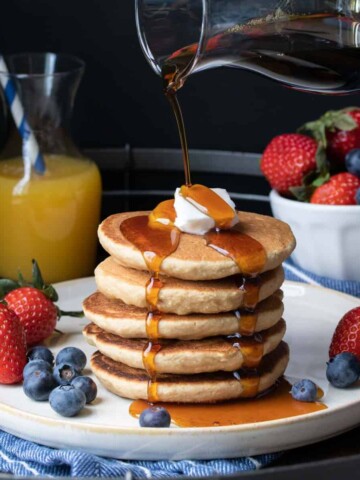 A glass container pouring maple syrup over a stack of pancakes and fruit