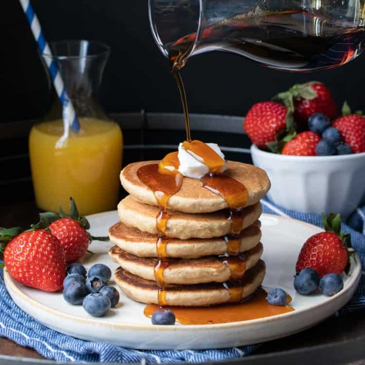 A glass container pouring maple syrup over a stack of pancakes and fruit