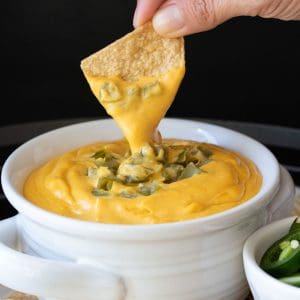 A tortilla chip dipping in a bowl of cheese sauce with chopped green chilis on top.