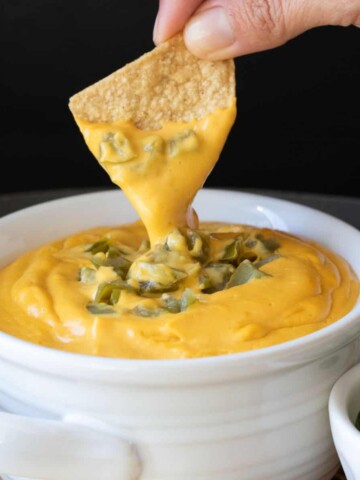 A tortilla chip dipping in a bowl of cheese sauce with chopped green chilis on top.