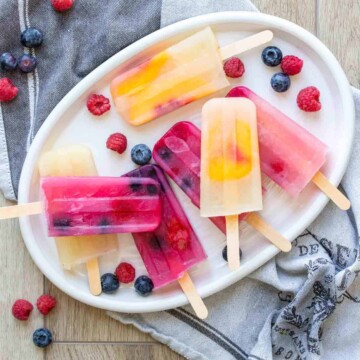 White platter with orange yellow and pink popsicles on it surrounded by berries