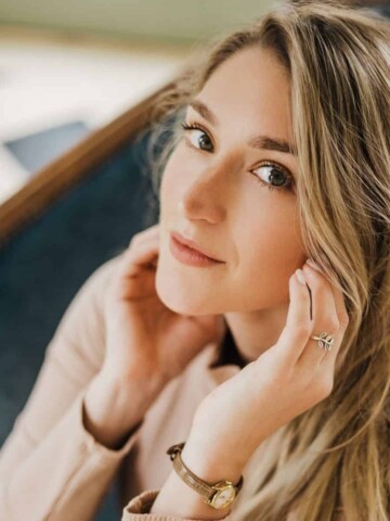 A woman with long blond hair and a slight smile with hands behind her ears