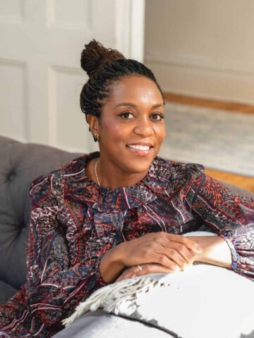 A black woman in a purple dress sitting on a couch with her hands folded on the couch arm