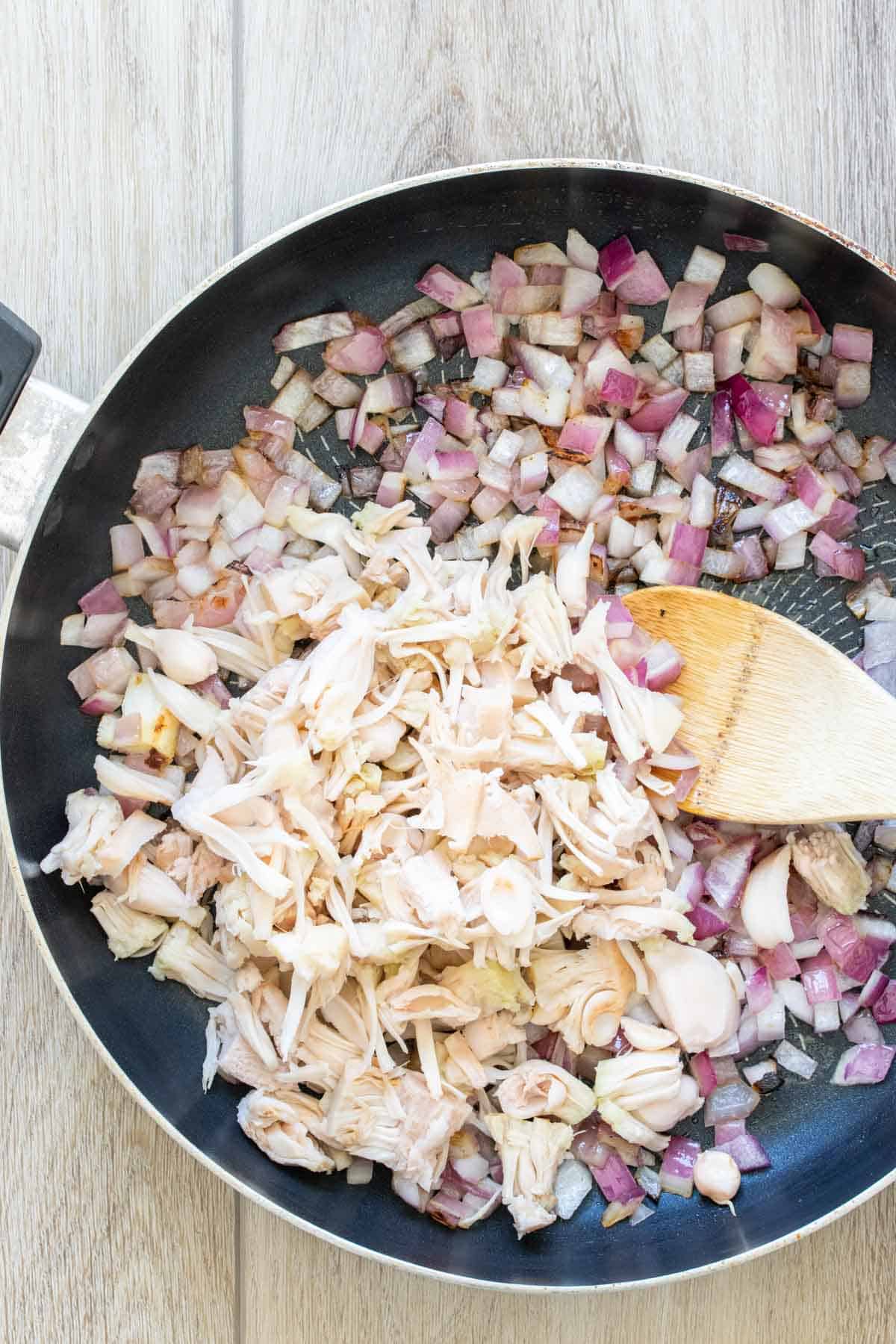 Jackfruit and red onion being mixed by a wooden spoon in a pan