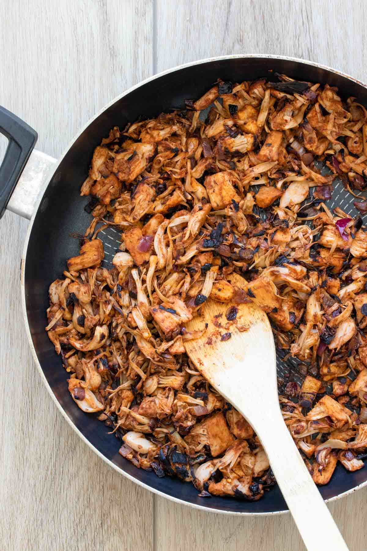 Wooden spoon mixing jackfruit with taco seasoning in a pan.