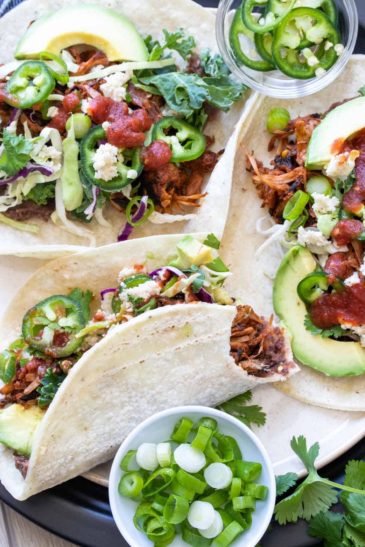 Jackfruit tacos on a plate with lots of toppings and one with a bite out of it.