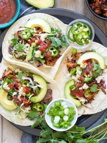 Three jackfruit tacos with lots of toppings on a black plate.