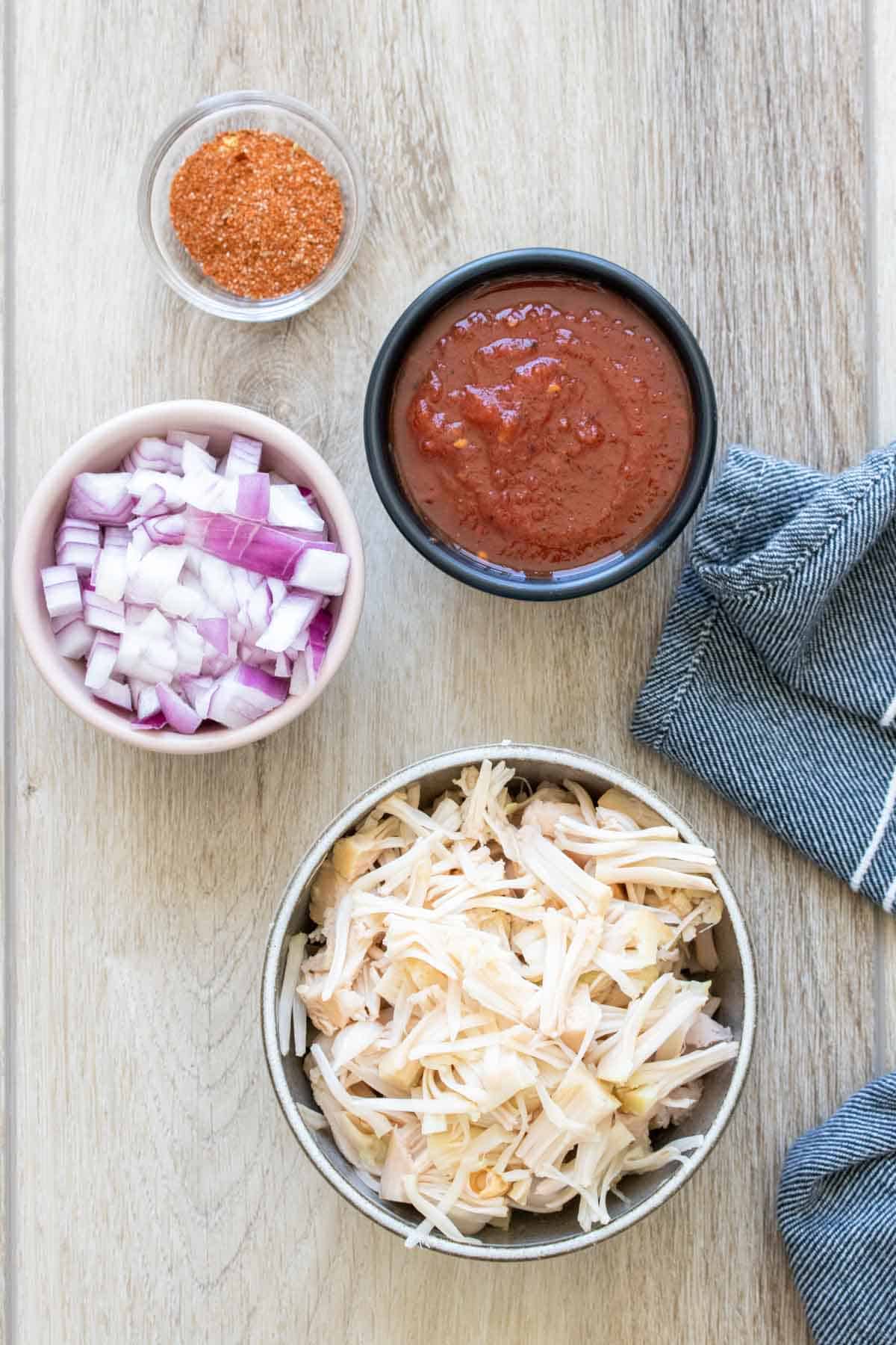 Four bowls with jackfruit, salsa, red onion and taco seasoning on a light wood surface next to a blue towel.