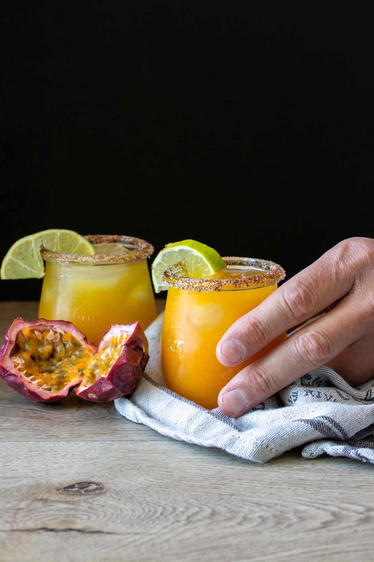 A hand grabbing a glass with an orange drink next to a passion fruit