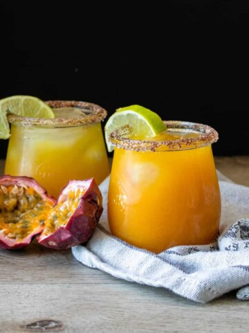 A cut open passion fruit next to two glasses with orange drinks