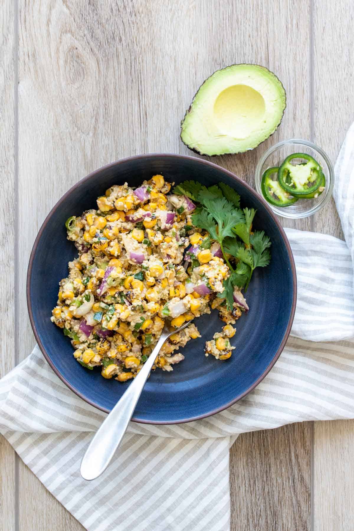 Corn salad with red onion and cilantro in a blue bowl next to avocado and jalapenos.