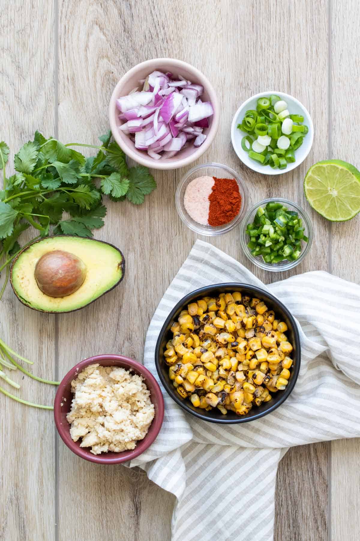 A tan wooden surface with bowls of ingredients needed to make an elote flavored corn salad.