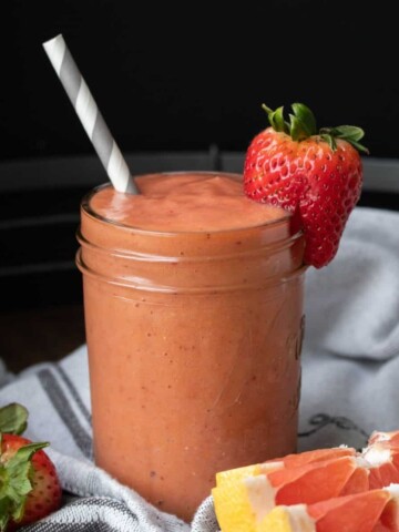 A jar with an orange smoothie in it and a grey and white straw next to fruit