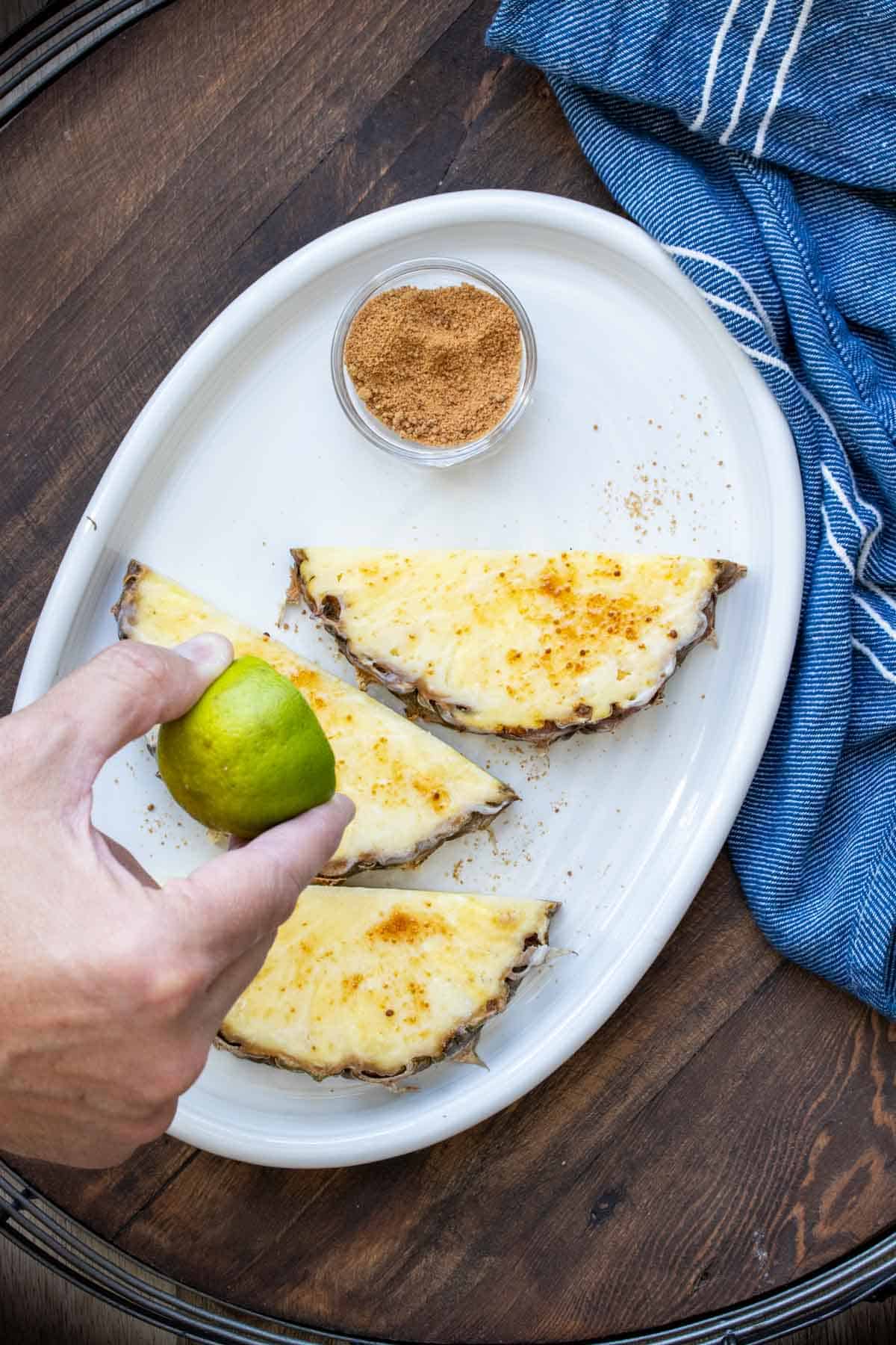 Hand squeezing a lime over sliced of pineapple on a white plate.