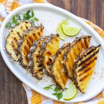 Slices of pineapple with grill marks on a white platter sitting on a yellow checked napkin