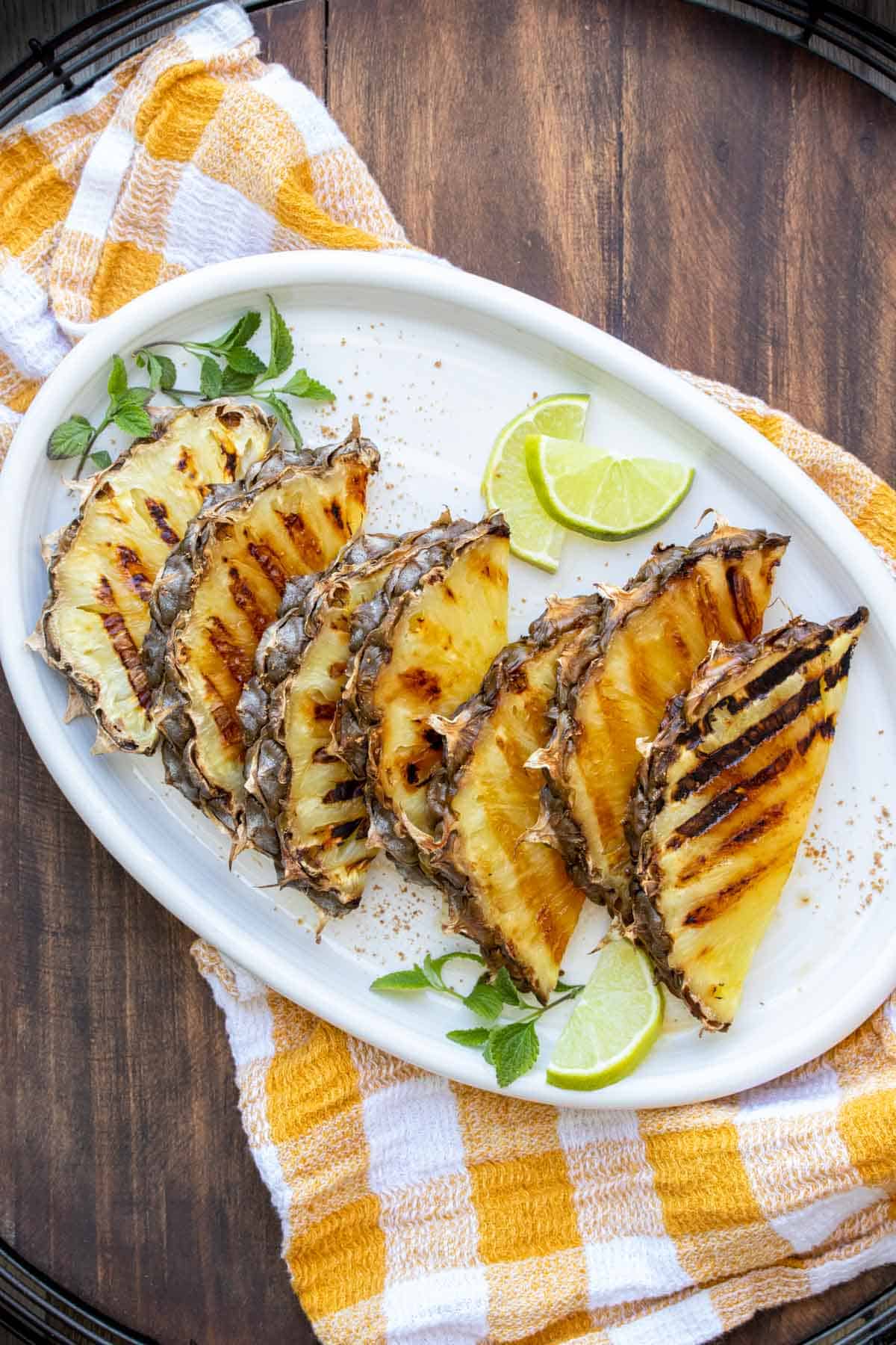 Slices of pineapple with grill marks on a white platter sitting on a yellow checked napkin.