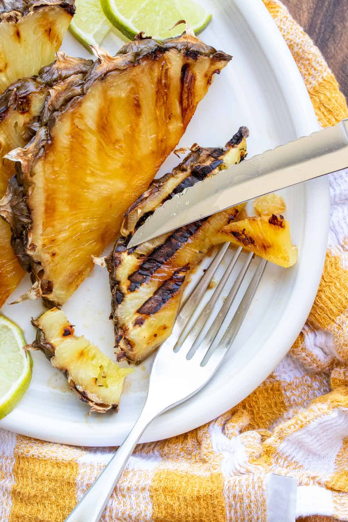 Fork and knife cutting a slice of grilled pineapple on a white plate.