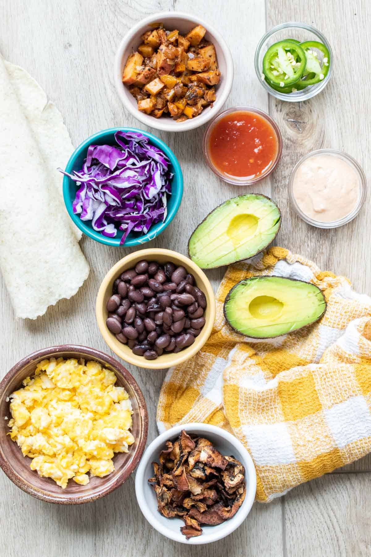 Bowls with ingredients to make breakfast burritos on a wooden surface and yellow checkered towel