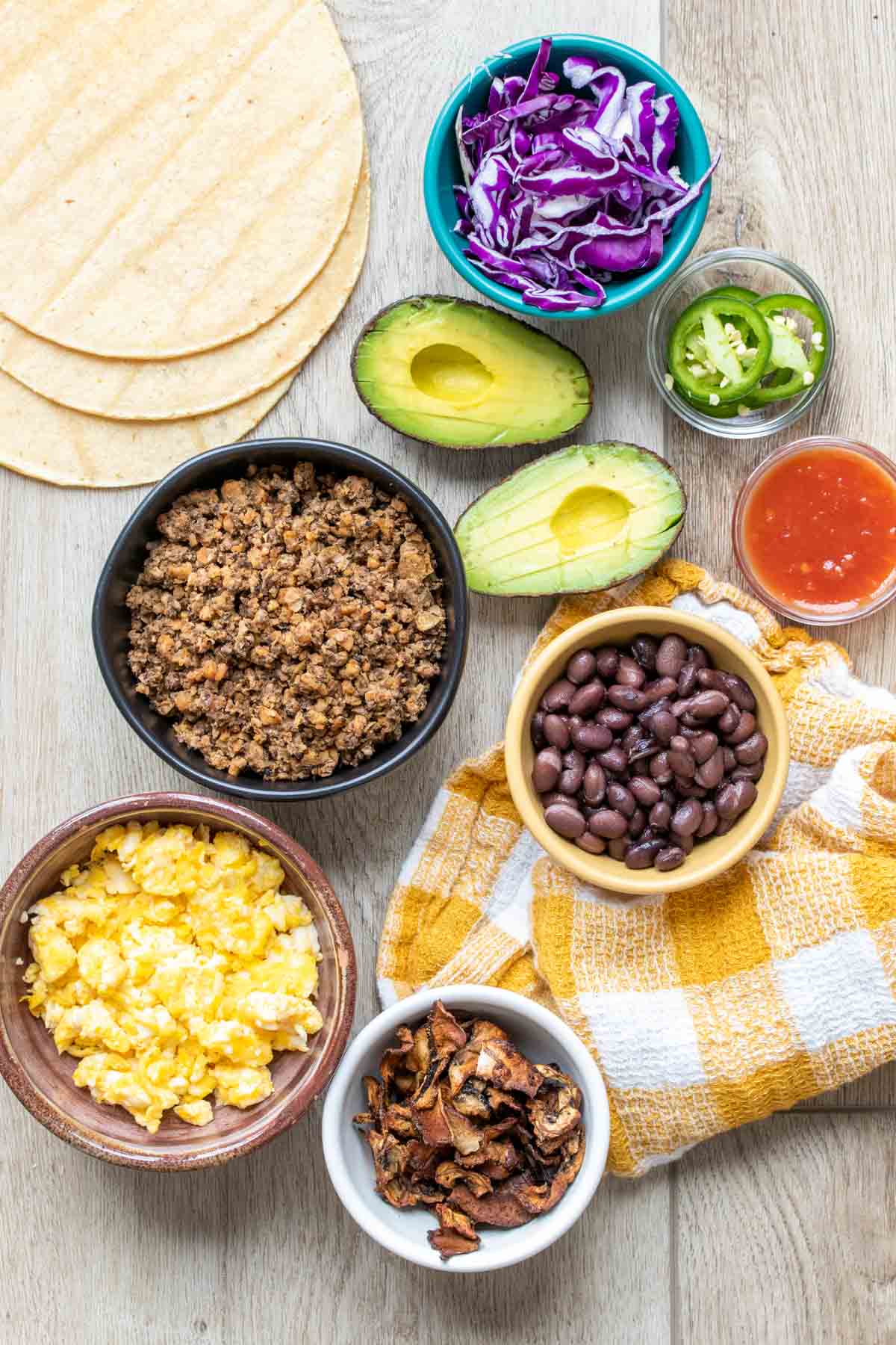 Ingredients in bowls needed to make breakfast tacos next to tortillas and avocado halves