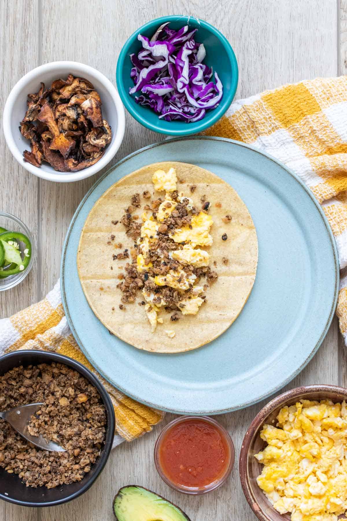 A blue plate with a tortilla on it being filled with things to make a breakfast taco