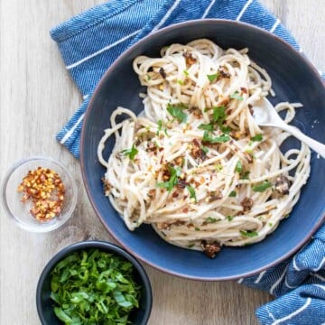 A blue bowl of spaghetti carbonara next to small bowls of parsley and red pepper flakes