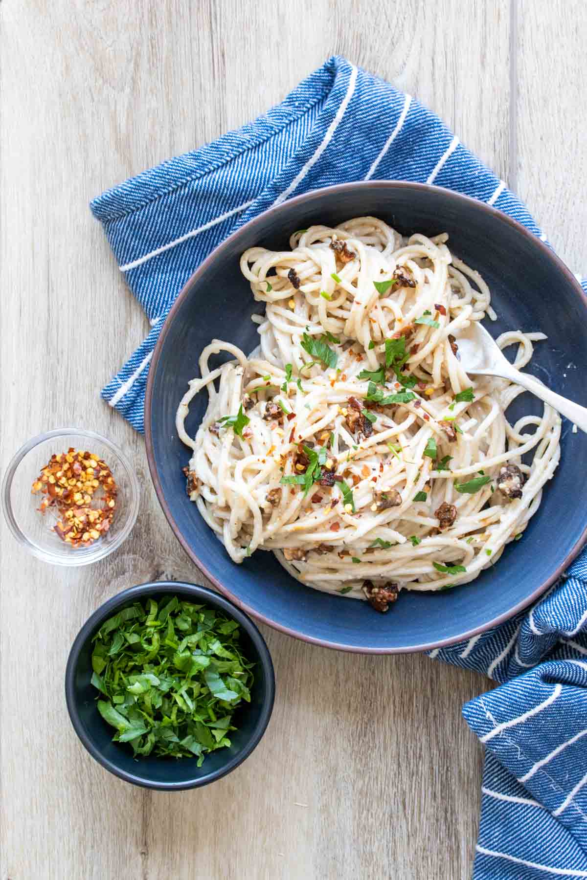 A blue bowl of spaghetti carbonara next to small bowls of parsley and red pepper flakes